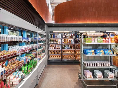 Modern convenience store interior with a large curved Corten steel wall, refrigerated display cases on the left, and industrial metal shelving units displaying liquor, snacks, and daily goods.