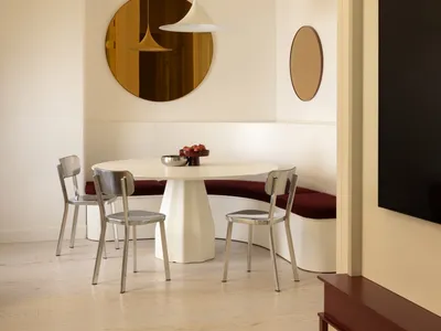 Modern dining area featuring a curved white banquette with deep red upholstery, a central white pedestal table, brushed steel chairs, a large round amber mirror, and a white pendant lamp.