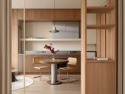 Modern minimalist dining area featuring a round wooden table, two cane and metal chairs, warm oak cabinetry, and a stainless steel pendant light.