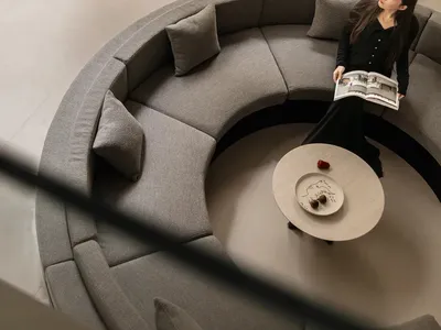 High-angle view of a modern living space featuring a large curved grey boucle sectional sofa surrounding a round coffee table, with a woman reading a book, opening to a lush sunken courtyard planted with moss, rocks and a Japanese maple tree.