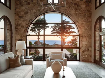 Luxurious living room featuring a massive arched picture window with ocean view at sunset, stone walls, cream sofa, and modern chandelier.