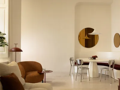 Modern dining area with a curved white pedestal table, built-in burgundy banquette, chrome chairs, a large round brass mirror, and a burnt orange boucle armchair in the foreground.