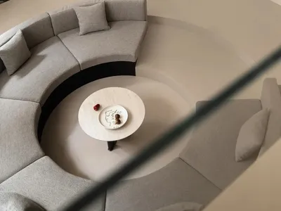 High-angle view of a modern living room featuring a large curved grey boucle sofa arranged around a round marble coffee table, with an indoor garden of ferns and rocks in the background and a black pendant lamp hanging above.