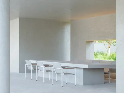 Minimalist concrete dining room featuring a long built-in concrete table, white metal-framed chairs, and a large horizontal window overlooking a green garden.