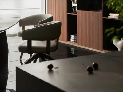Modern dining area featuring two beige upholstered swivel chairs, a dark dining table, and a tall walnut built-in shelving unit displaying art objects, with lush green plants in the foreground.