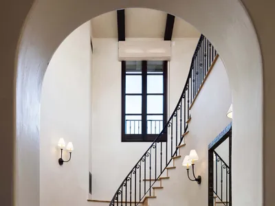 View through a large arched doorway into a spacious entryway featuring a curved wooden staircase with ornate black wrought-iron railing, terracotta tile floor, dark wood console table, and wall sconces.