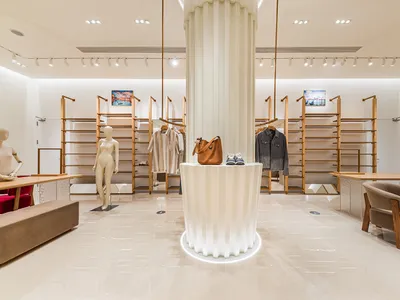 現代極簡服裝精品店內部，中央有大型白色凹槽立柱展示台、木製服裝展示架、人體模特兒，以及米色布面扶手椅，搭配拋光大理石地板。