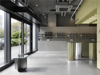Modern minimalist retail interior with polished concrete floors, textured concrete walls, cylindrical stainless steel counter, and a bright lime green perforated tile wall with integrated shelving.