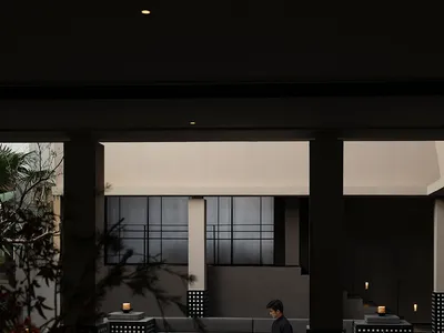 Night view of a modern minimalist concrete courtyard with built-in grey sofas, a man sitting in the sunken lounge area, warm candlelight, and dark architectural pillars.