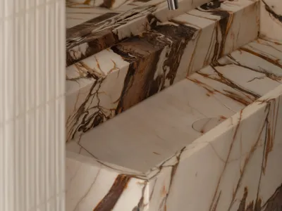 Close-up of a luxurious Calacatta marble vanity sink with dramatic brown veining and a modern brushed nickel wall-mounted faucet against a neutral textured wall.