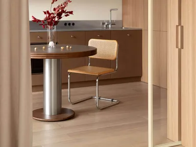 View of a modern minimalist kitchen dining area with walnut cabinetry, a round wooden dining table on a metal pedestal, a cane and leather Cesca-style chair, stainless steel countertop and sink, and a large metallic pendant lamp.