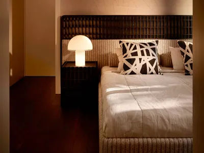 Modern minimalist bedroom with dark wood grid headboard, textured beige wallpaper with geometric line pattern, white mushroom table lamp, and black and white abstract pillows on a woven bed base.