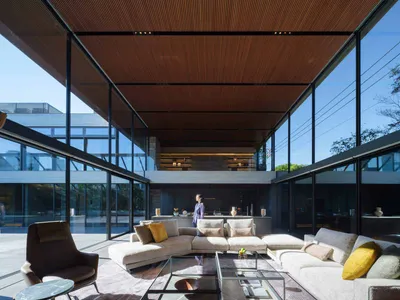 Modern double-height living room featuring a large L-shaped sectional sofa, brown leather armchair, glass coffee tables, and dramatic wood slat ceiling with full-height glass walls overlooking a courtyard.