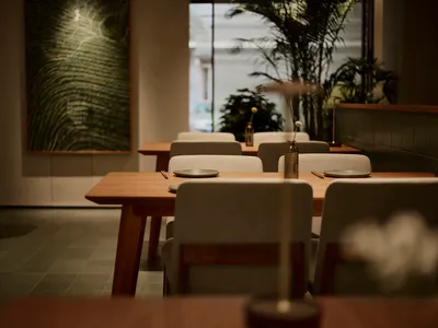 現代簡約餐廳用餐區，設有淺色橡木餐桌、米色布面座椅、牆上大型綠色紋理藝術畫作，以及靠近大窗的豐富室內植物。