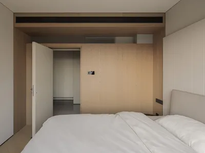 現代極簡臥室，設有大型淺橡木嵌入式衣櫃、布面床頭板、白色寢具，以及通往走廊的開放門口。