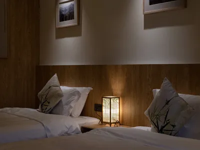Cozy twin beds in a Japanese-inspired bedroom featuring vertical wood paneling, white bedding with bamboo-print pillows, and a lit rectangular lantern on a wooden nightstand.