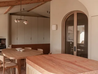 Modern minimalist dining room featuring a large oval oak dining table, wooden chairs, a terracotta stone kitchen island in the foreground, built-in banquette seating, exposed wooden ceiling beams, and a fireplace in the background.