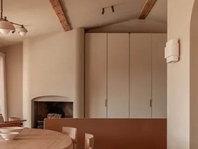 Warm minimalist dining room with round oak table, wooden chairs, terracotta floor tiles, and a fireplace in the background under exposed wooden ceiling beams.