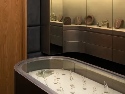 Interior view of a luxury jewelry store featuring curved walnut wood cabinetry, dark padded wall panels, and illuminated glass display cases with gold jewelry.