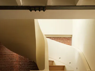 View of a modern curved wooden staircase with light-colored plaster walls and an exposed red brick wall in the background.