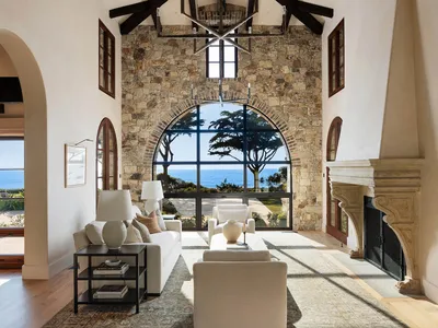 Spacious coastal living room featuring a large stone fireplace, white sofas, high beamed ceilings, and a dramatic arched picture window overlooking the ocean and trees.