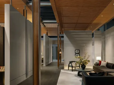 Modern open-plan living area featuring concrete walls, wooden ceiling slats, a dark sectional sofa, and a large floral arrangement on a round coffee table.