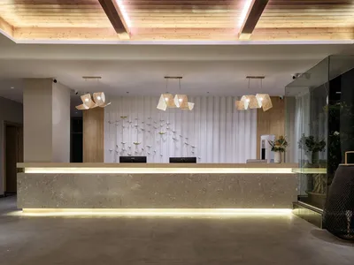 Modern hotel reception desk made of travertine stone with under-lighting, woven rattan pendant lamps, white pleated wall, metallic bird wall art, and wooden ceiling.