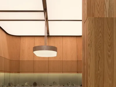 Interior of a high-end jewelry boutique featuring curved oak paneling, dark green marble floor tiles, a cylindrical dark metal display island, and a large circular pendant light.