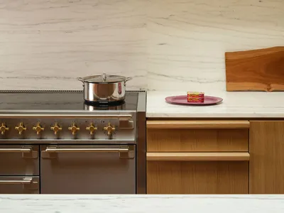Modern kitchen corner featuring a stainless steel range with brass knobs, white marble backsplash and countertop, light oak cabinetry, and a wooden cutting board with colorful cookbooks on an upper shelf.