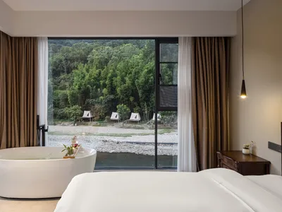 Modern hotel bedroom featuring a large white freestanding bathtub positioned directly in front of a floor-to-ceiling window overlooking a lush green forest and river, with a wooden four-poster bed and dark wood nightstand.