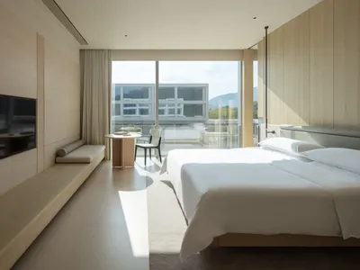 現代簡約飯店臥室，淺橡木板壁、大型平台床鋪白色寢具、嵌入式窗邊座椅，以及落地玻璃門外壯麗的山景。