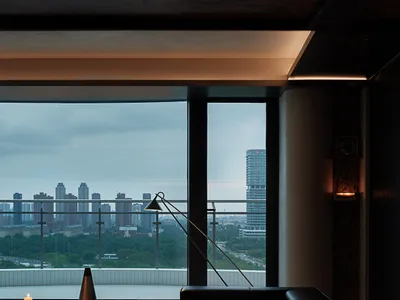 Modern living room interior at night featuring a black leather sofa, multiple lit candles, minimalist dark wood furniture, and large floor-to-ceiling windows overlooking a city skyline with buildings and greenery.