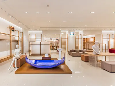 Modern fashion boutique interior featuring a glossy blue Venetian gondola display on a wooden platform, surrounded by minimalist oak shelving and white mannequins under bright track lighting.