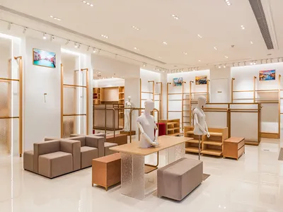Modern minimalist fashion boutique interior with beige modular sofas, light oak display tables with textured glass bases, white mannequins, brass shelving units, and glossy white tiled floors.