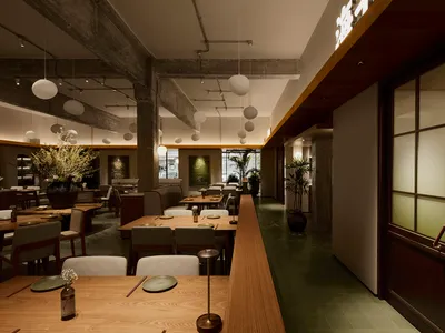 Interior view of a modern Japanese restaurant featuring long light oak dining tables, upholstered chairs, exposed concrete beams, and hanging spherical pendant lights.