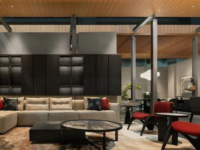 Modern lounge area with a large beige sectional sofa, dark walnut wall unit, concrete accent wall, round marble coffee table, and red accent chairs under a warm wood-paneled ceiling.