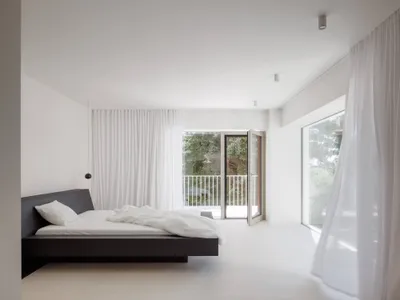 Modern minimalist bedroom with a dark wood floating platform bed, white bedding, sheer white curtains, and large glass doors opening to a balcony with greenery.
