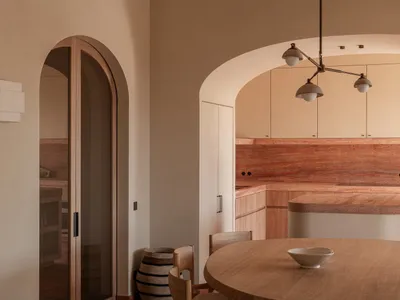 Warm minimalist kitchen and dining area featuring rounded wooden dining table, light oak cabinetry, terracotta marble backsplash, and dramatic arched doorways under a modern multi-arm pendant light.