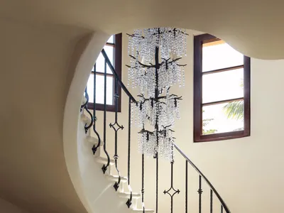 Elegant curved staircase with black wrought-iron railing, large crystal chandelier, and round limestone table with decorative vase and candles in a bright entryway.