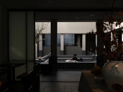 一位男子坐在現代極簡日式住宅的混凝土下沉式中庭休息區，從深色用餐區望去，前景有花器。