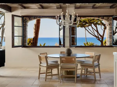 Round dining table with four cane-back chairs positioned in front of large arched windows opening to a bright blue ocean view, white chandelier hanging from beamed ceiling, limestone tile floor.