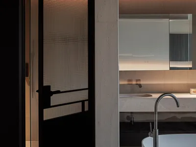 Modern minimalist bathroom featuring raw concrete walls, a white freestanding bathtub with a curved floor-mounted faucet, a stone floating vanity, and a black-framed ribbed glass door with warm backlighting.