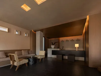 Modern minimalist lounge and reception area featuring exposed concrete walls, dark tiled floor, built-in beige sofa, woven cane armchairs, and a black reception counter with warm recessed lighting.