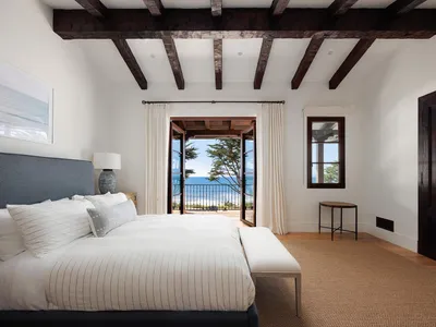 Bright coastal bedroom with large bed dressed in white striped linens, dark wood ceiling beams, and open French doors revealing a stunning ocean view.