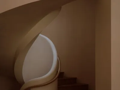View of a modern curved staircase featuring a smooth white plaster balustrade, warm wooden handrail, and dark walnut treads ascending into soft beige walls.