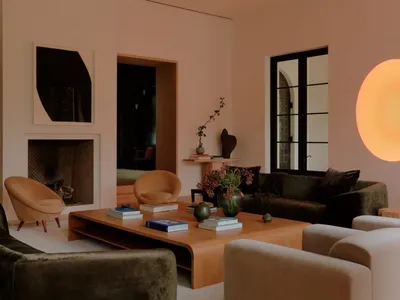 Modern living room featuring dark green velvet sofas, tan curved armchairs, a large oak coffee table, minimalist fireplace, and large spherical wall light.