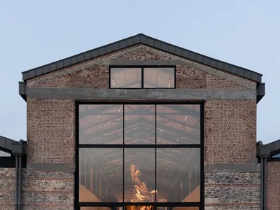 Front exterior view of a converted industrial brick building featuring a massive black-framed glass facade revealing the brightly lit interior gallery space at dusk.