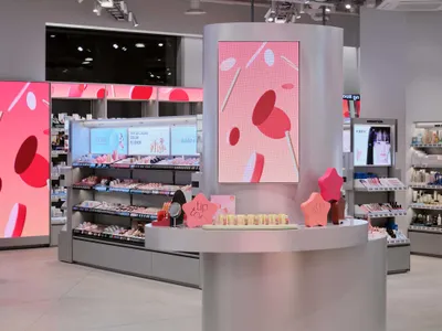 Modern silver curved beauty counter kiosk with large pink digital screen and product displays in a brightly lit retail space