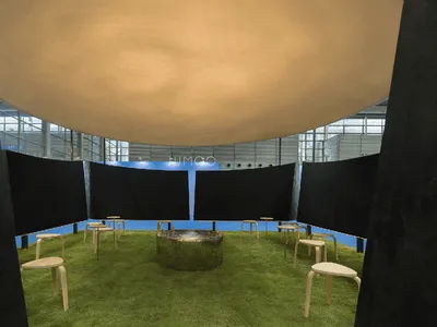 Modern art installation showing a large beige fabric dome suspended above a circular green artificial grass floor with wooden stools and a central metallic fire pit.