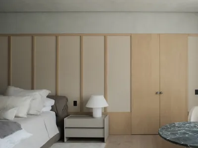 Modern minimalist bedroom with light oak built-in wardrobe, beige upholstered headboard, white bedding, and a round green marble table in the foreground.